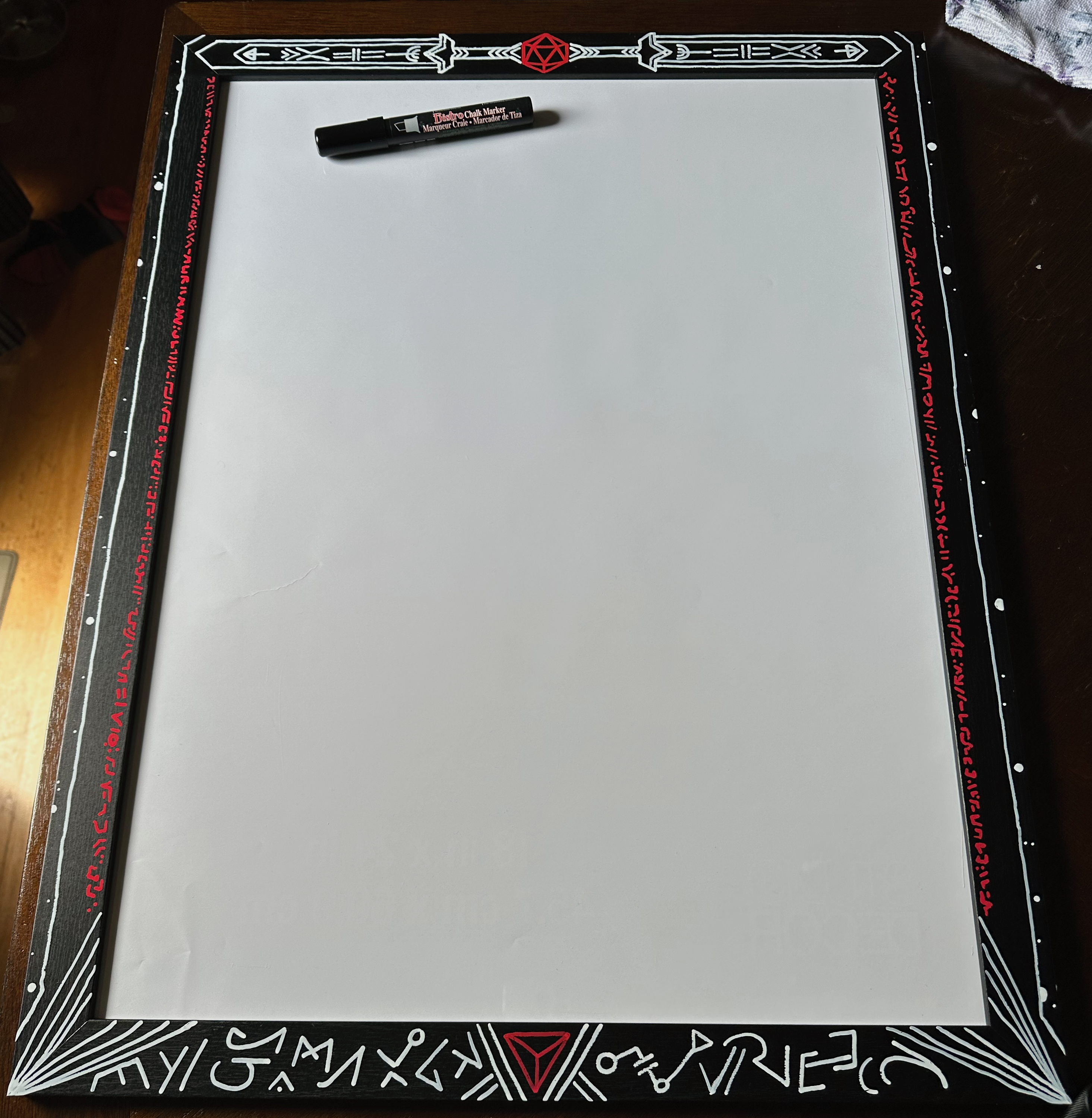 A black frame decorated with white and red runes, patterns, swords, and dice. It’s framing an empty white space with a black chalk marker lying on top.